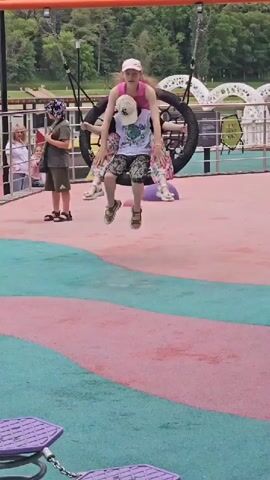 A group of children and an adult are visible at a playground. One child is on a tire swing with another child sitting on their lap. An adult stands nearby, arms crossed. The playground has a pink and green surface with colorful climbing structures and swings. In the background, there is a body of water and trees.
