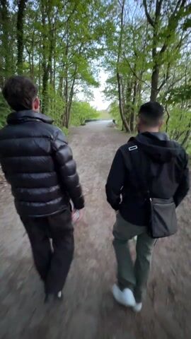 Two young men walk away from the camera along a dirt path through a dense forest with tall trees, their branches forming a green canopy overhead. The path leads to a slight uphill area. The man on the left is wearing a shiny black puffer jacket and dark trousers, carrying a small item in his hand. The man on the right is wearing a hooded jacket and green cargo pants, with a small black messenger bag slung across his body. The light suggests it is late afternoon or early evening.
