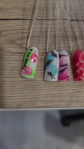 The video shows a fan of nail swatches displayed on a wooden surface. The swatches showcase a variety of nail art designs, including floral patterns, abstract swirls, animal prints like zebra and leopard, polka dots, a palm tree silhouette, a skull, balloons, and a pink pattern with the word "FOREVER". The lighting appears to be overhead, casting subtle shadows.