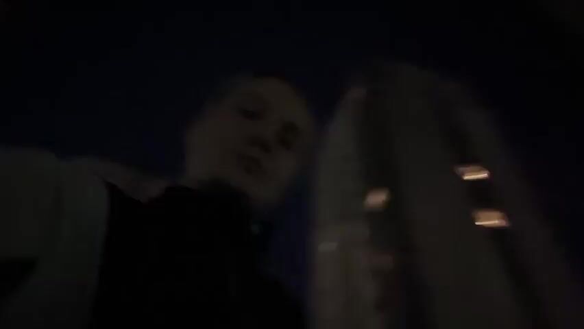 The video, shot at night, features a person with short hair, wearing a black turtleneck and a light-colored jacket. The camera angle is low, looking up at the person. The background shows a tall building with lit windows. The camera moves slightly as the person walks or shifts position. The lighting is poor, making the scene dark and difficult to see details clearly.