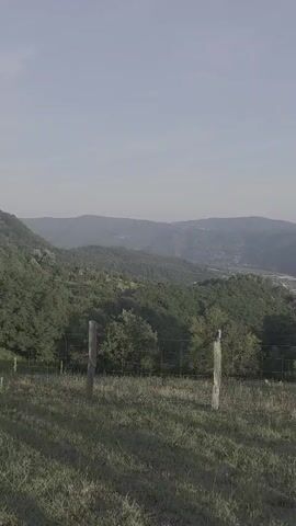 This is a vertical, wide shot of a rural landscape in Mali Zvornik Municipality, Serbia. The foreground features a grassy field with a wire fence supported by wooden posts. The middle ground consists of rolling, tree-covered hills, and in the distance, a valley with a village and a body of water can be seen. The sky is overcast and hazy, suggesting a cloudy or misty day. The overall mood is peaceful and serene, with a sense of vastness and natural beauty. There is no visible text in the media file.