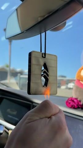 A person's hand holds a lighter, igniting the edge of a wooden air freshener hanging from a car's rearview mirror. The air freshener is rectangular with a cutout of a surfboard. The background shows a bright blue sky and a blurred outdoor environment.