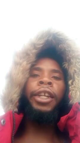 A man with dark skin, a beard, and a fur-lined hood is speaking directly into the camera. He is wearing a red jacket. The background is bright, likely an overcast sky. He mentions showing viewers fishermen fishing.