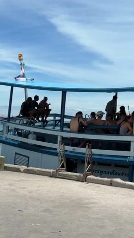 The video shows a boat docked at a pier. There are many people on the boat, some sitting and some standing. A woman in a pink shirt and black shorts is climbing onto the boat, carrying a large bag. Another person in a grey shirt and backpack is already on the boat. In the background, there are several other boats and a blue sky with white clouds. The pier appears to be made of concrete, and there are some bags and luggage lying on the ground.