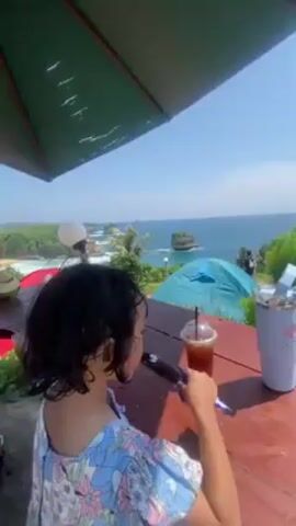 A girl with dark, curly hair sits at a wooden table, eating a chocolate bar and holding a drink. She is wearing a blue and white floral shirt. On the table, there is a clear plastic cup with a dark liquid and a straw, and a silver cup containing ice. In the background, there is a scenic view of the ocean and a coastline with trees and rocky outcroppings. There are two tents pitched on the land near the ocean. A green umbrella partially covers the scene from above. The sky is clear and blue.