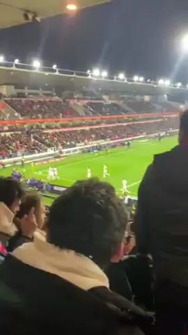 From a seated position in a stadium, the video shows a section of the audience watching a soccer match. The stadium is packed, and the field is brightly lit. The teams are visible on the field, wearing white and dark jerseys. The crowd is cheering, and the atmosphere appears to be energetic. The perspective is from behind a spectator, who has dark curly hair and is wearing a light-colored hoodie under a black jacket.