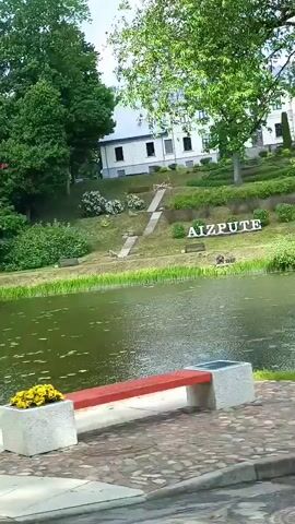 The video displays a still shot of a scenic outdoor environment. In the foreground, a tranquil body of water, likely a pond or lake, is visible, partially covered with floating green vegetation. Beyond the water, a grassy slope rises, featuring large white block letters spelling out "AIZPUTE." To the left of the letters, a set of outdoor stairs ascends the slope. Further in the background, a large, multi-story building with a light-colored facade and numerous windows is situated amidst abundant green trees. The sky above appears overcast. A small section of a paved surface and a dark pole are visible in the bottom left corner. No people or animals are present in the frame.