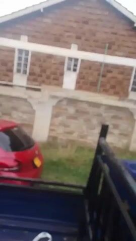 The video shows a blurry view of a neighborhood from the back of a moving blue vehicle, likely a pickup truck or similar type of vehicle. There are houses with brick walls and white trim visible, as well as trees and patches of grass. Other cars, like a red one, can be seen parked along the street. There's also a utility pole with visible wiring. The video is shaky and out of focus, likely due to the movement of the vehicle.