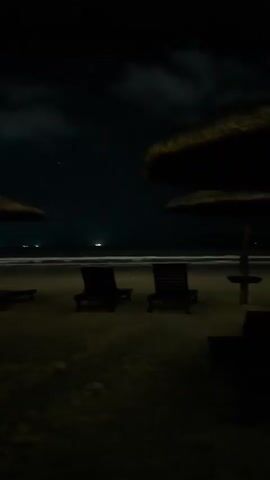 A dark beach at night. Several empty lounge chairs and thatched umbrellas are visible on the sand. The ocean is visible in the background with gentle waves and distant lights. The sky is dark with some clouds. The scene is still and quiet.