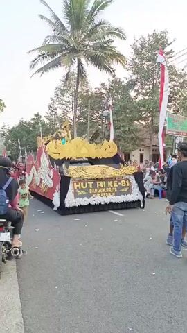 A parade float adorned with a golden dragon and the text "HUT RI KE-81 BANGUN GIRIYA SUMBEREJO" moves down a street lined with trees and spectators. The float is decorated with white and red accents. A young child in a green shirt runs in front of the float. Several people are standing and watching the parade.