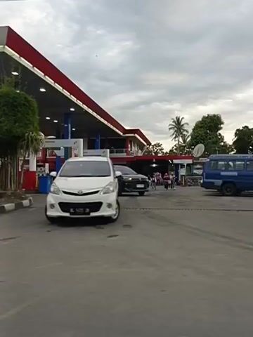 The video shows a gas station with several cars and motorcycles. The gas station appears to be a Pertamina gas station, indicated by the signage. The weather is cloudy. In the foreground, a white car is driving away from the gas station. In the background, there are several other vehicles, including a red car and a white van, as well as some trees and buildings. A person on a motorcycle is also visible. The video is shot from a moving vehicle.