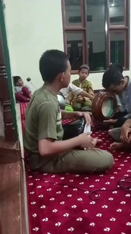 A group of young boys are sitting on a red carpet with a white star pattern, engaged in playing traditional musical instruments. The boys are wearing Islamic attire. In the foreground, one boy in a green shirt is playing a black drum. To his right, another boy in a green shirt and black skullcap is holding a drum. Behind them, a boy in a light-colored shirt is holding a round drum and appears to be playing it. Further back, another boy in a patterned shirt is seated and looking towards the front. In the background, a man is visible. The setting appears to be an indoor space with wooden window frames.