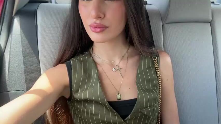 This is a close-up selfie video of a young woman with long, dark hair, sitting in the back seat of a car. She is wearing a green pinstripe vest over a black top and several gold necklaces, including one with a cross pendant and another with a rectangular tag. She has on light makeup and is looking directly at the camera with a neutral expression. The background shows the gray interior of the car, including the headrest and part of the window. The lighting suggests it is daytime, possibly late morning or early afternoon, with bright sunlight filtering in. The overall scene is casual and personal, likely a candid moment captured while traveling. There is no visible text in the video. The location is Aventura, United States of America.