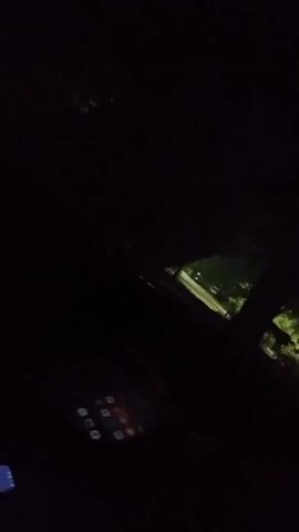 The video begins with a dark frame, then an illuminated outdoor scene appears. A streetlight casts light on palm trees and foliage. A paved path or road is visible, with two figures walking in the distance. The scene is dark and appears to be at night. The camera angle is tilted.