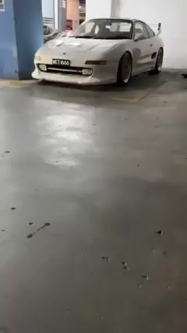 A white, modified Toyota MR2 is parked in an indoor parking garage. The car has a wide body kit, gold wheels, and a rear wing. The license plate reads "MET 1666".  The garage floor is grey concrete with yellow painted lines. A blue column is on the left side of the frame. Someone can be heard saying "Bye".