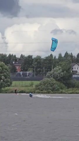 A video shows a person kitesurfing on a body of water. The kitesurfer is in the foreground, riding on the water's surface and kicking up spray. In the background, there are trees and buildings, with a cloudy sky overhead. Several kites are visible in the sky, one in the foreground and two further back. One kite is blue, another is black and white, and the third is purple and blue. The person in the foreground is wearing a blue and white suit. The scene appears to be a recreational area.