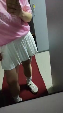 This is a selfie video taken by a person in what appears to be a locker room or dressing area. The person is wearing a pink t-shirt with cartoon characters on the left chest and white text across the front. They are also wearing a white pleated skirt, white socks, and light-colored sneakers. The person has light brown hair and is looking at the phone while filming. The background shows lockers and a red mat on the floor. The text on the shirt reads "WE ARE THE FUTURE". The person is adjusting their hair.