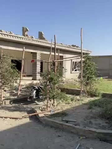 The video shows a man standing in front of a damaged building. The building appears to have been damaged by an explosion or some other type of destructive event. There is debris scattered all around the building. The man is wearing a black suit and a green headscarf. He is looking at the camera and talking. The sky is blue and the sun is shining.