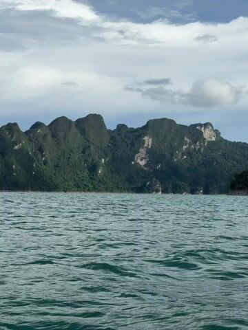 This media file is a short video clip showing a scenic view of Khao Phang, Thailand. The primary subjects are the majestic limestone karsts covered in lush green vegetation that rise dramatically from a vast body of calm, turquoise water. The scene is set on a body of water, likely a lake or a wide river, with the mountains forming a stunning backdrop.

The activity taking place is a boat ride, as indicated by the gentle rocking motion of the water and the perspective from a moving vessel. The water is slightly rippled, suggesting a light breeze. The weather appears to be overcast with scattered clouds, creating a soft, diffused light across the landscape. The time of day is not explicitly stated but the lighting suggests it could be late morning or afternoon. The overall mood conveyed is one of tranquility and natural beauty, with the imposing mountains evoking a sense of awe and wonder. There is no visible text in the media file.