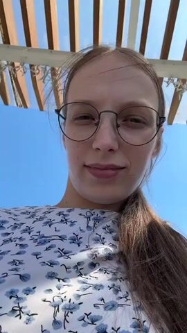 A young woman with long brown hair and glasses smiles into the camera. She is wearing a white dress with a blue floral pattern. The video appears to have been shot outdoors on a sunny day, with a bright blue sky visible in the background. Some wooden structures are visible overhead. The woman is touching her hair.