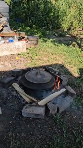 A black, lidded pot sits on a fire made of wood and bricks in an outdoor setting. Flames and smoke rise from the fire. Several pieces of wood are placed around the pot. In the background, there is green foliage and a white surface with some bottles and other objects on it. The ground is covered in dirt and grass. The sun is shining, casting shadows.
