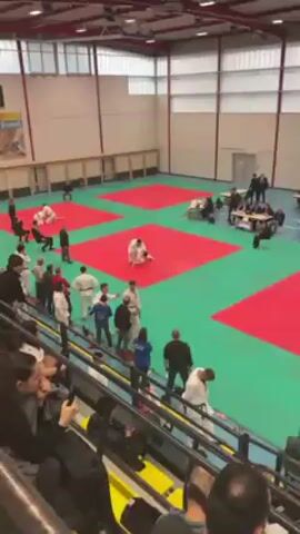 The video shows a judo competition taking place on a green mat with red squares marked out. Several people wearing judo uniforms are present, with two actively engaged in a match. One judoka executes a throw, causing his opponent to fall onto the mat. Several other people in judo uniforms are standing around the mat, observing the match. Other individuals, some in casual clothing and one in a suit, are also watching from the sidelines. The event appears to be held indoors, possibly in a gymnasium or sports hall, with additional spectators seated in the background.