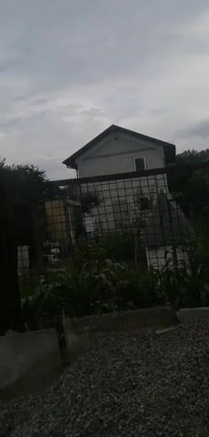 A gray sky looms over a residential area. A two-story house with a dark roof stands in the background, partially obscured by a chain-link fence. The fence encloses a yard filled with lush green plants and some white structures. In the foreground, a pile of gray gravel and concrete blocks creates a rough texture. The scene appears to be a cloudy day in a somewhat overgrown or unkempt outdoor setting.