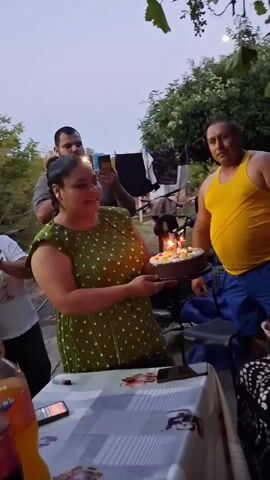 The video shows a gathering of people outdoors, possibly for a birthday celebration. A woman in a green polka-dot dress holds a chocolate cake with lit candles. She is smiling and looking towards the cake. To her right, a man in a yellow tank top and blue shorts stands behind her, also looking at the cake. To the left of the woman, a man in a plaid shirt and another person partially visible are present. In the background, there are trees and foliage, suggesting an outdoor setting. The lighting appears to be dim, possibly evening or dusk. A table with drinks and a phone is visible in the foreground. The atmosphere seems festive.