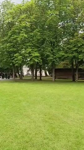 The video pans across a grassy lawn in front of a traditional wooden and white-washed building with a wooden roof. Several people are seated at picnic tables on the lawn in front of the building, some engaged in conversation. Trees and foliage surround the building and the lawn. In the background, a group of people are gathered, and some vehicles are visible. The sky appears overcast. The camera movement is a steady pan from left to right.
