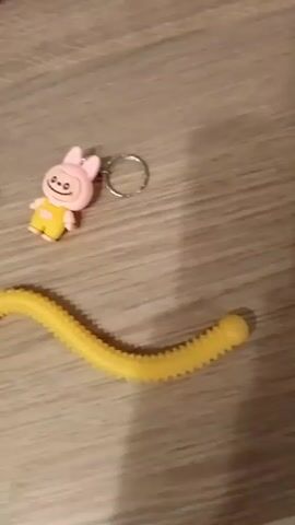 On a light brown, wood-grained surface, there are three keychains and a yellow corrugated tube. The tube is curved and positioned horizontally across the lower part of the frame. The keychains are arranged above the tube. On the left is a pink keychain with green ears. In the center is a similar pink keychain with a rabbit-like face and wearing yellow clothing. A third keychain is visible, partially out of frame on the left side.