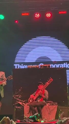A live musical performance is taking place on a dimly lit stage. In the foreground, a person is seated and playing a sitar. To their left, another person stands and plays a red electric guitar. Both musicians appear to be focused on their instruments. Behind them, a large screen displays a graphic resembling a stylized, concentric semi-circle pattern, with text overlayed that reads "Thievish Corporation". Stage lighting, predominantly red and green, illuminates the scene with beams cutting through smoke or haze. The overall atmosphere suggests a concert or live event.