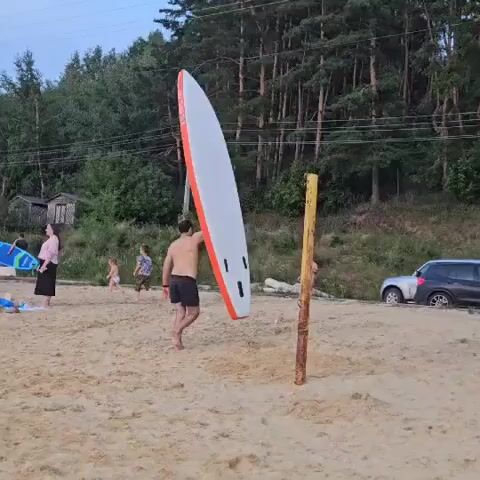 A man is standing on a sandy beach, holding a white surfboard with orange trim upright next to a wooden pole. He is wearing black shorts and is shirtless. In the background, there are trees, a car stuck in the sand with its trunk open, and a few other people. The sky is overcast.