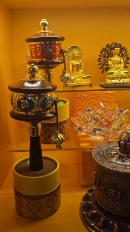 A close-up shot displays a collection of Tibetan Buddhist artifacts on orange shelves. In the foreground, two large, ornate prayer wheels stand prominently. The prayer wheels are made of dark metal with intricate carvings and symbols. Smaller decorative items, including crystal lotus flowers and what appear to be smaller prayer wheels or decorative boxes, are placed on the shelves behind the larger prayer wheels. In the background, several golden Buddha statues of varying sizes are displayed on the top shelf, some adorned with halos and set against decorative backdrops. The lighting is warm, casting a golden glow over the entire scene, suggesting an indoor setting like a shop or a shrine. The camera slowly pans across the display, offering a detailed view of each artifact.