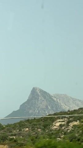The video shows a mountain landscape from a moving vehicle, likely a car, as indicated by the streaks and smudges on the glass in the foreground. The sky is a hazy light blue. In the distance, a prominent rocky mountain with a pointed peak is visible. In the middle ground, there is a rocky, green, and shrub-covered hillside with a white barrier or railing running across it. The vegetation consists of dry grasses, bushes, and some scattered rocks. The motion of the vehicle causes the foreground elements to blur, creating a sense of movement through the landscape. The overall impression is a scenic drive through a mountainous, possibly Mediterranean, environment.