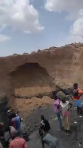 The video shows a group of people working or gathering in a dirt pit or excavation site. There are several individuals, some using tools that appear to be shovels or hoes, digging into the dark, muddy ground. Others are standing around, observing or resting. The excavation is against a backdrop of a large dirt wall, possibly a bank or cliff face, with visible layers of soil. The sky above is partly cloudy, with patches of blue visible between the white clouds. The overall setting seems to be in a dry, possibly rural environment.
