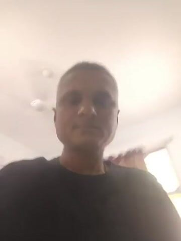 A medium-skinned, short-haired man wearing a black t-shirt faces the camera. The shot is from a low angle, looking up at the man. The background consists of white walls and a white ceiling with a ceiling fan. The man looks around, moving his head slightly. The camera is moving.