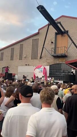 This media file captures a lively outdoor concert scene in Stockholm, Sweden. A band performs on a stage in front of a large brick building with large windows and a red awning. The building's facade is decorated with vertical slats and a prominent black metal structure extends from the roof. A white circular backdrop with colorful drawings and pink drapes serves as the stage's focal point.

A crowd of people, dressed casually in summer attire, gathers to watch the performance. The atmosphere appears energetic and joyful, with many individuals visible from behind, facing the stage. Some people are wearing hats, including a straw hat and baseball caps. The band members, including a vocalist, guitarist, and drummer, are actively engaged in playing music.

The time of day appears to be late afternoon or early evening, as indicated by the warm, soft lighting and the sky, which shows a mix of clouds and dusky hues. The weather seems pleasant, suitable for an outdoor event. The overall mood is celebratory and engaging, suggesting a positive audience experience. There is no discernible text visible in the media.