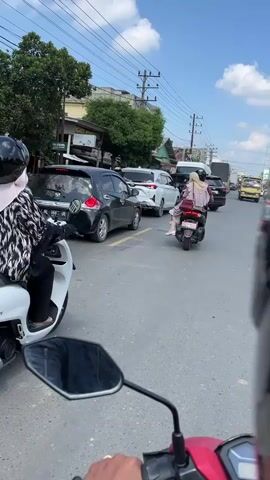 The video shows a first-person perspective of someone riding a motorcycle in a traffic jam. Several other motorcycles and cars are visible, as well as a few pedestrians and buildings. The sky is blue with some clouds, and the weather appears to be warm. The rider's hands are visible on the handlebars of the motorcycle, and a helmet is on their head. The overall impression is of a busy urban street scene.