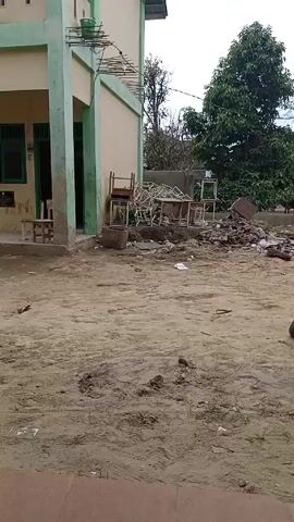 The video shows a two-story building with green and beige paint. In front of the building is a sandy area with scattered debris, including broken chairs, tables, and other discarded items. A large tree is visible in the background on the right side of the frame. The sky is cloudy. A voice can be heard in the background.