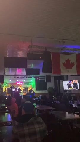 A vertical video shows a live band performing on a stage in a dimly lit venue. The band consists of four male musicians playing guitars and singing. The stage is lit with colorful stage lights, including pink, green, and blue. Above the stage, a large Canadian flag is prominently displayed. A Cuban flag is also visible hanging on the wall behind the stage. In front of the stage, an audience is gathered, some standing and some seated at tables. The overall atmosphere appears to be a concert or a lively gathering.