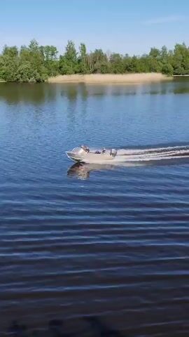 The video captures a small motorboat traversing a wide, calm body of water under a clear blue sky. The boat, light in color, is equipped with an outboard motor that propels it, creating a distinct white wake in its trail. At least two individuals are visible in the boat as it moves from the left side of the frame towards the right.

In the background, a distant shoreline features a dense line of green trees and what appear to be reeds or tall grasses, suggesting a natural, unurbanized setting. The water itself is a deep blue, reflecting the bright sunlight, and shows subtle ripples on its surface. The weather appears fair and sunny, indicative of a pleasant day for boating. The activity suggests leisure and enjoyment of the outdoors. The scene takes place in Chaika, Russia. No visible text is present in the video.