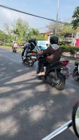 The video shows a street scene with several motorcycles in motion. The camera perspective appears to be from another vehicle, possibly a motorcycle as well, positioned behind and slightly to the side of the subjects. The foreground is dominated by the asphalt road surface, marked with shadows from overhead trees. In the mid-ground, two individuals are riding motorcycles, both wearing helmets. The rider in the foreground is wearing a brown hoodie and dark pants, astride a black scooter. Behind them, another person on a similar scooter is wearing a blue jacket and also has a helmet. Further back, another motorcycle with a rider is visible, and in the far distance, a white van is seen. The environment appears to be an urban or suburban setting, with trees lining the side of the road and buildings visible in the background. The lighting suggests it is daytime, with bright sunlight casting distinct shadows.