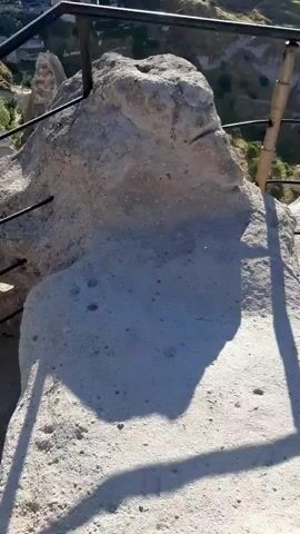 The video begins with a close-up of a large, weathered rock formation, possibly part of a cliff face. A metal railing runs horizontally in front of the rock, suggesting a viewing platform or walkway. The camera then pans upward and to the right, revealing a panoramic view of a valley filled with unique geological formations, resembling fairy chimneys. In the distance, a town or village is visible, with buildings nestled into the hillsides and a sprawling landscape stretching to the horizon under a clear sky. The overall scene is one of a natural wonder combined with human settlement.