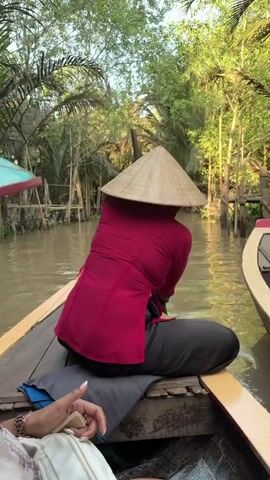This video captures a tranquil boat ride through a narrow, tree-lined canal, likely in the Mekong Delta region of Vietnam, given the context of Thoi Son.

The primary subject is a woman seated in the stern of a wooden sampan, facing away from the camera. She wears a vibrant red long-sleeved top and a traditional Vietnamese conical straw hat (nón lá), which obscures her face. She is actively paddling the boat through the murky green water.

In the foreground, a passenger is partially visible, facing slightly towards the camera. She is smiling subtly, wearing sunglasses, and a unique light blue conical hat with a colorful tropical pattern. Her right hand, adorned with rings and a bracelet, holds a green smartphone, suggesting she might be documenting the experience.

The setting is characterized by dense, lush tropical vegetation, including palm trees and various other green plants, that line both sides of the canal. Simple, rustic wooden structures, such as posts and what appear to be small jetties or bridges, are visible along the banks, blending with the natural environment. The scene is well-lit by natural daylight, indicating a pleasant, possibly sunny, day. The boat glides smoothly through the water, with the paddle creating gentle ripples, conveying a sense of peaceful movement and immersion in nature. The traditional attire and riverine landscape are strong cultural indicators of the location.