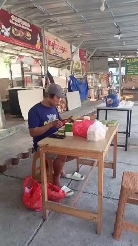 A man in a blue t-shirt and a grey cap sits at a wooden table outdoors, facing right and cutting small green cakes with a knife. He is barefoot and wearing white sandals. In front of him on the table are several stacks of green cakes, a red plastic bag, and white packaging. Two red plastic bags are on the ground to his left, and another is on the ground to his right. He is using a small cutting board. The background shows a covered outdoor area with various food stalls. One stall sign reads "Angkringan". Another stall has a sign with "WARU" and some green text. There are other tables and chairs visible. The ground is concrete.