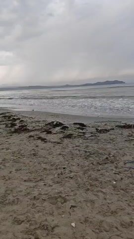 The video is an outdoor shot of a beach with a grey, overcast sky. The foreground is dominated by sand with some debris like seaweed and small objects. In the mid-ground, the ocean has moderate waves rolling towards the shore. In the background, a distant landmass, possibly islands or a coastline, is visible under the clouds. The camera appears to be stationary, capturing the scene without any visible human presence or activity.