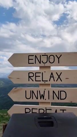 The video shows a wooden sign with four arrows pointing left, each inscribed with a word: ENJOY, RELAX, UNWIND, and REPEAT. Below the sign, a black object is visible. The camera slowly pans upward and to the left, revealing a vast landscape of rolling green hills and mountains in the distance, partially covered with snow. The sky is bright blue with scattered white clouds. The entire scene depicts a serene and natural environment.