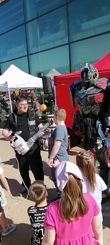 An outdoor scene features a man in a black vest with "SWAT" text holding a metallic, guitar-shaped object towards a boy in a grey t-shirt. To their right, a person in a detailed red, blue, and silver robot costume stands. Several children are in the foreground and background, observing; one girl in the immediate foreground has her arm raised. A white tent, a red canopy, a speaker on a stand, colorful bunting, and a large glass building are visible in the background under what appears to be a sunny sky.