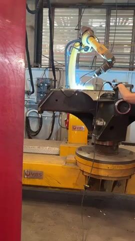 A robotic arm is welding a metal component. The robotic arm is yellow and has multiple joints. It is positioned above the metal component, which is black and appears to be part of a larger structure. A bright light emanates from the welding torch on the robotic arm. Sparks are visible. A person's hand is visible on the right side of the frame, near the metal component. The environment appears to be a factory or industrial setting, with exposed pipes and machinery visible in the background. The floor is dark and appears to be concrete. There are yellow and red painted surfaces in the scene.