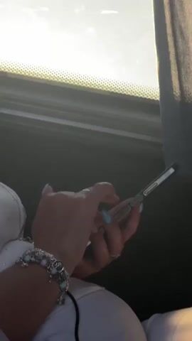 A person's hands and forearms are visible, wearing a white top and a charm bracelet. The person is holding a smartphone in their right hand and appears to be interacting with it, possibly typing or scrolling. The left hand is also near the phone, with a ring on one finger. The background shows a window with bright light filtering through, suggesting the scene takes place near a window, possibly in a vehicle or building. The overall lighting is somewhat dim, with sunlight creating a lens flare effect.